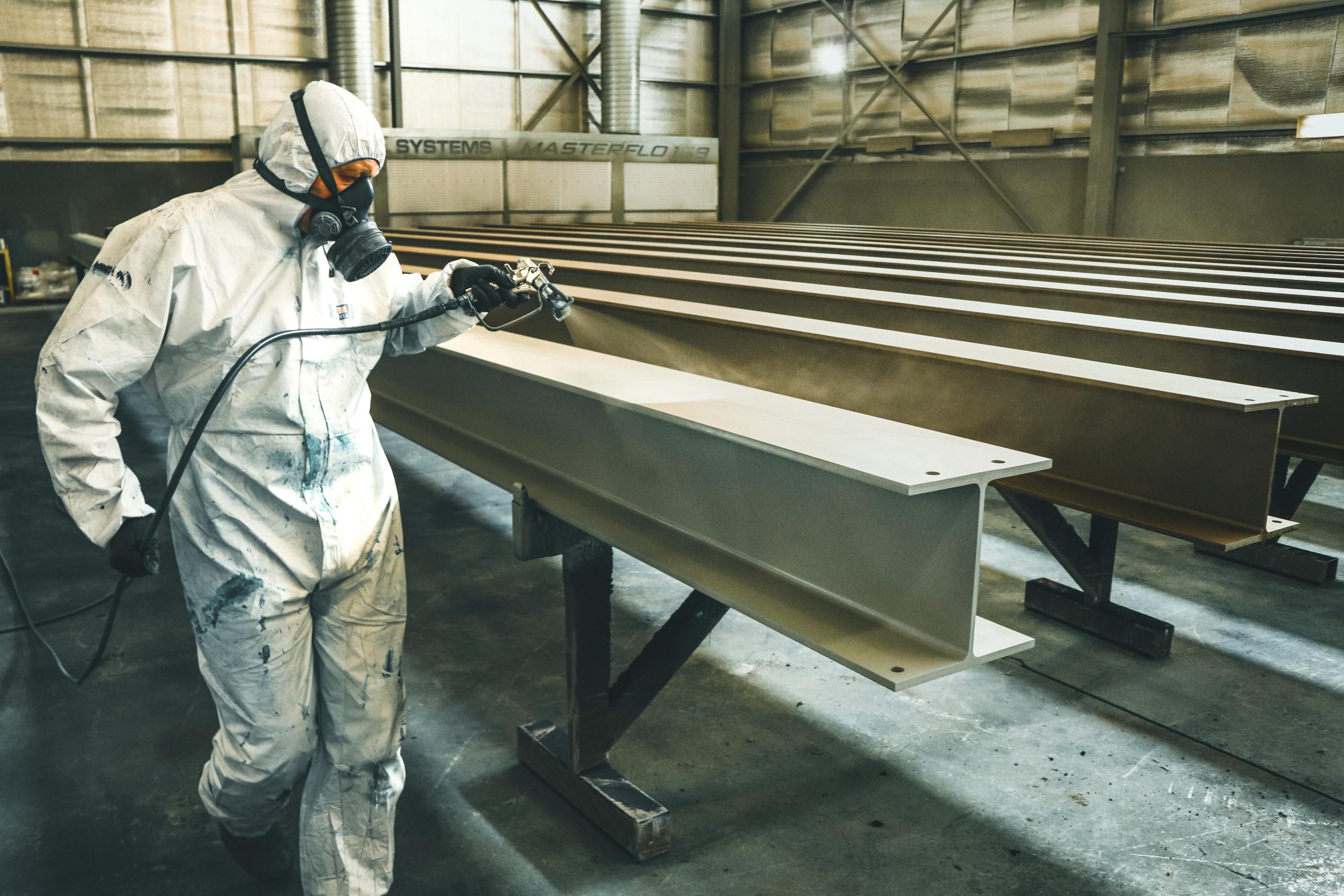 Worker in protective suit spray painting metal beams in an industrial setting indoors.